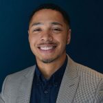 Issac Patterson-Ramsey, Corporate Social Responsibility Associate, SMBC smiles to camera in a suite against a navy blue background.